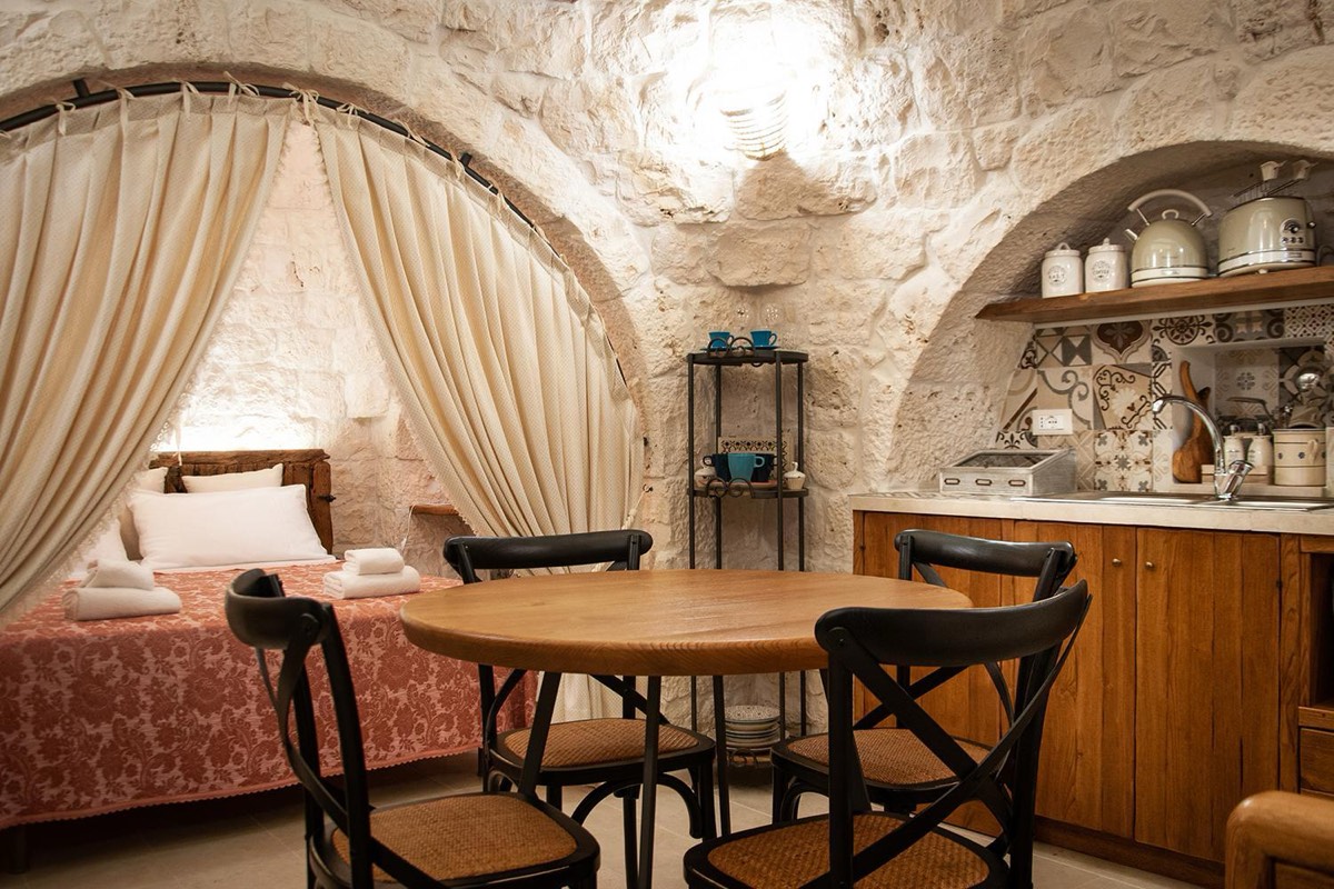 L’ambiente unico: il letto sotto un arco con le tende, il tavolo rotondo e l’angolo cottura
