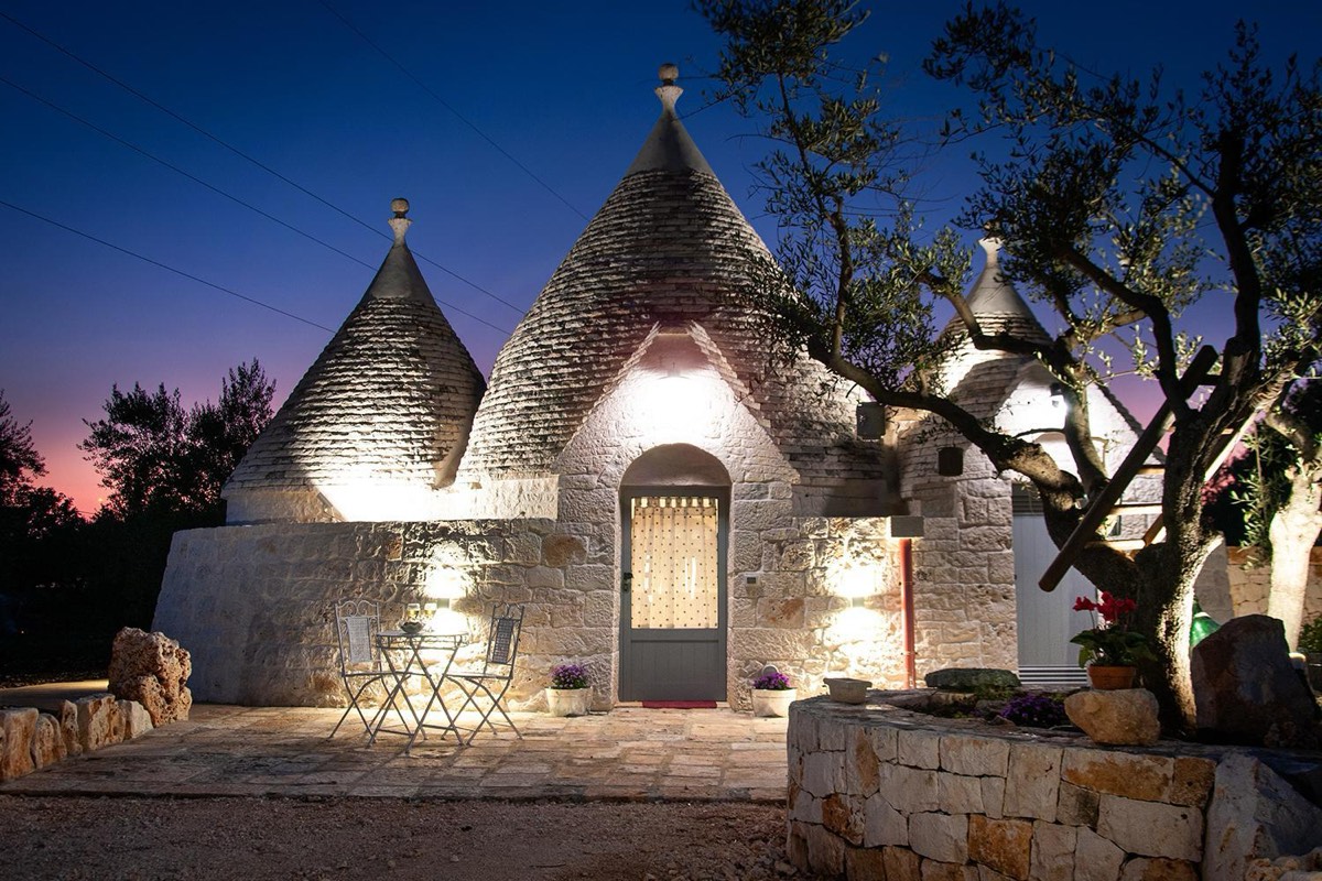 Il trullo all’imbrunire, con le luci accese sotto i coni e un ulivo accanto